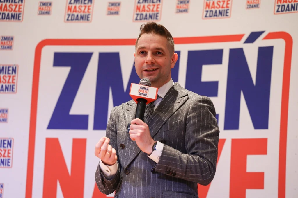 Patryk Jaki przemawiający z mikrofonem na tle logo Zmień Nasze Zdanie.