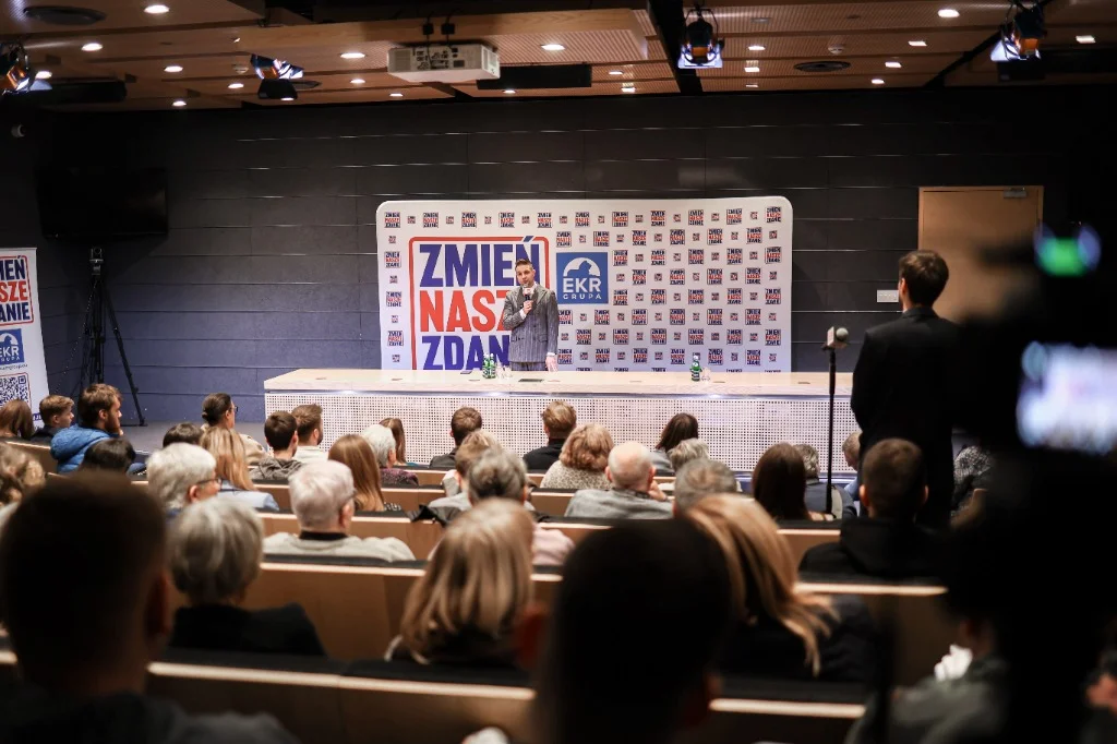 Widok auli uniwersyteckiej podczas debaty Zmień Nasze Zdanie z Patrykiem Jakim na scenie i kamerą na statywie.